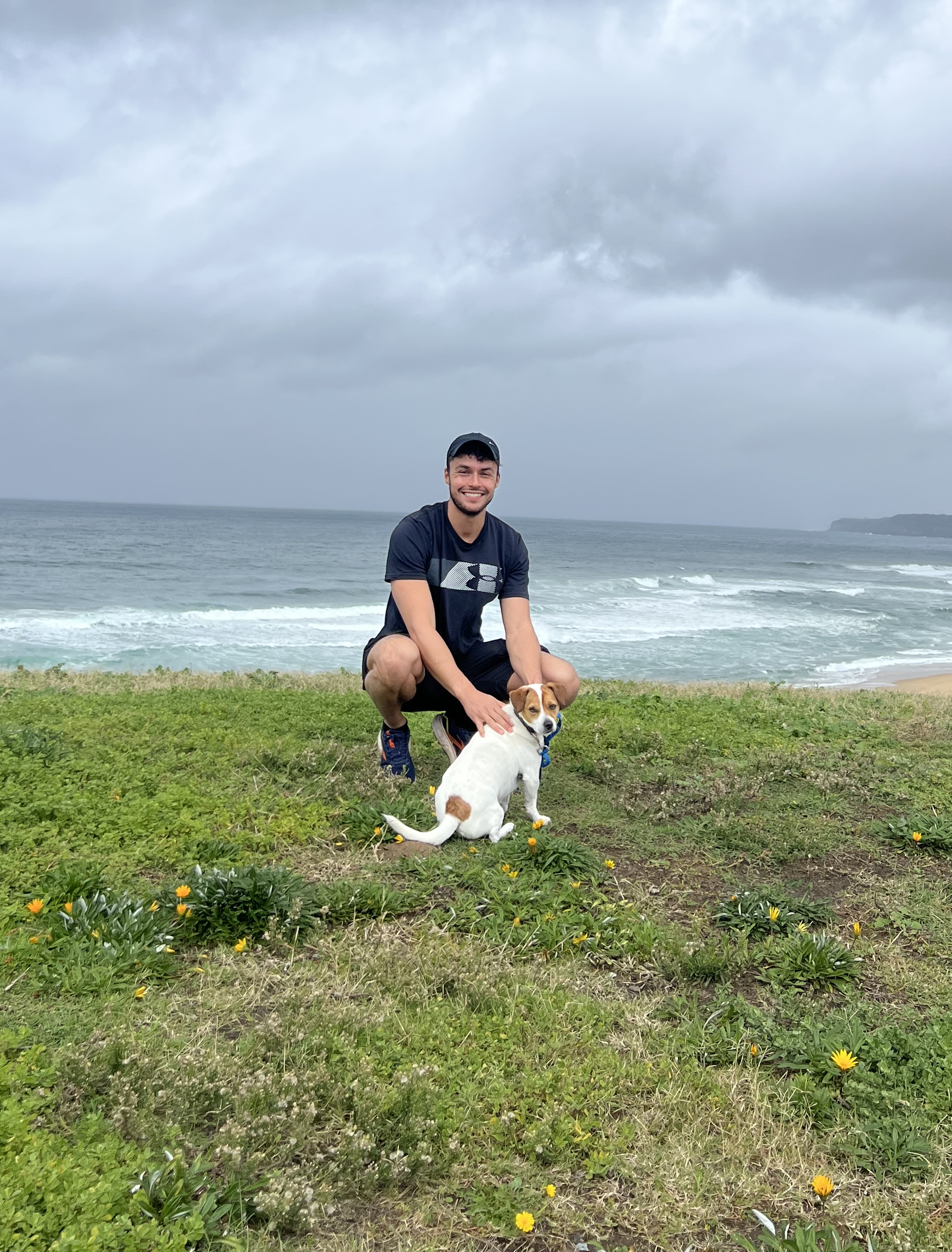 Man and dog sitting on a grassy hill by the ocean under a cloudy sky